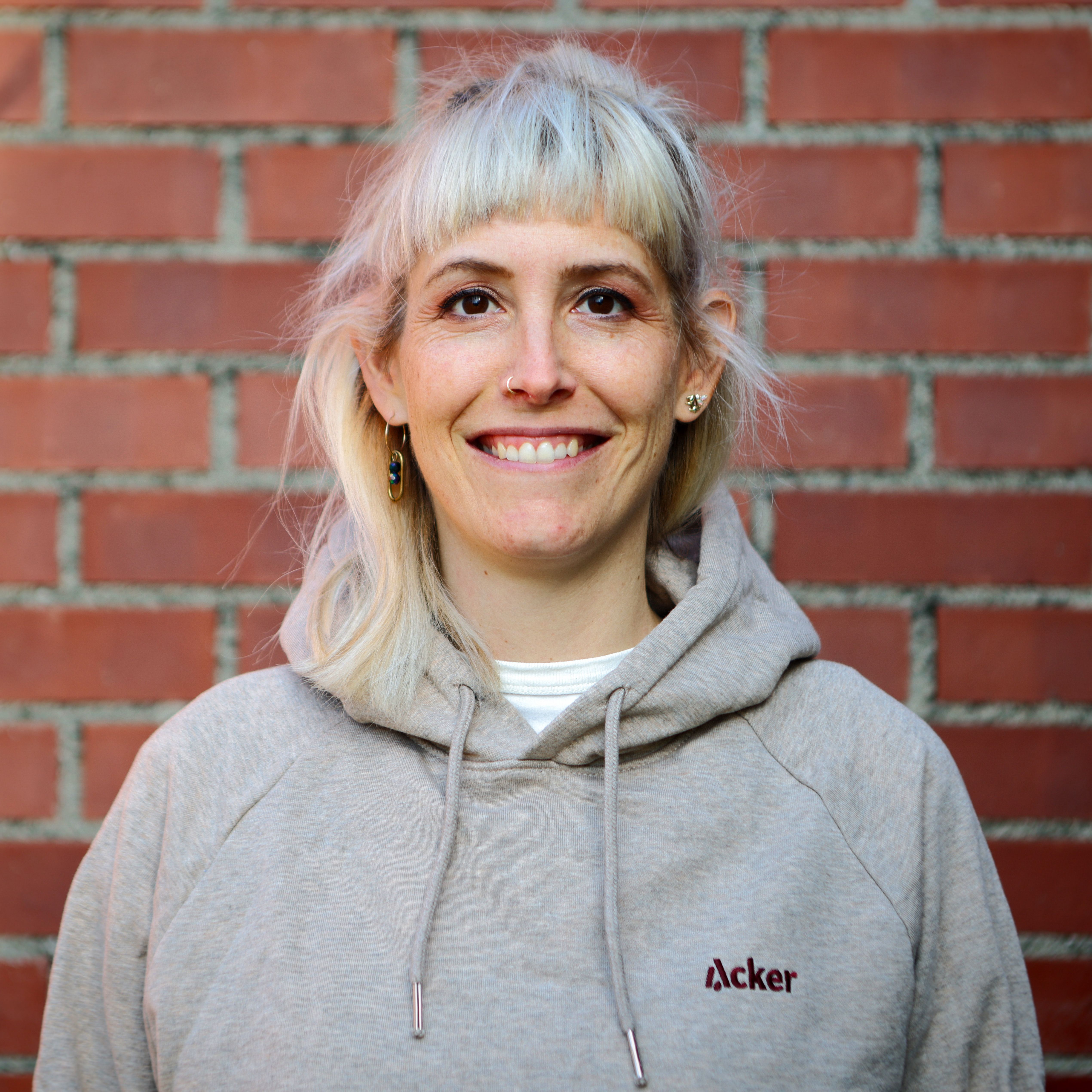 Porträtaufnahme von Simone Nägeli. Sie lächelt in die Kamera; grauer Hoodie mit «Acker»-Logo, Backsteinwand im Hintergrund.