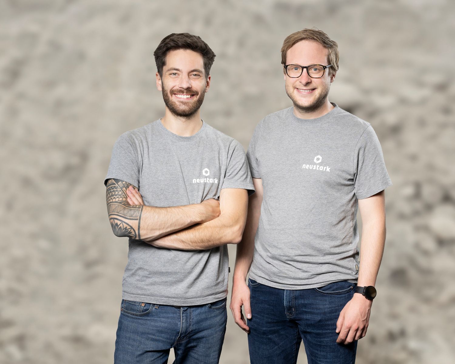 Die neustark-Mitgründer Valentin Gutknecht und Johannes Tiefenthaler stehen nebeneinander vor neutralem Hintergrund und tragen graue T-Shirts mit dem neustark-Logo.