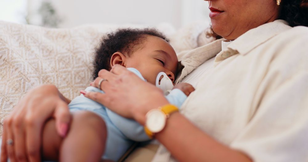 Eine Frau sitzt auf einem Sofa und hat ein Baby in einem Strampler auf der Brust, das schläft