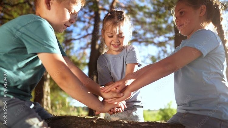 Drei Kinder stehen im Freien in einem Wald und legen ihre Hände übereinander, als Zeichen von Teamgeist und gemeinschaftlichem Spielen.