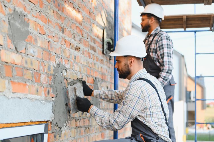 Zwei Handwerker mit Schutzhelmen verputzen auf einer Baustelle eine Backsteinwand.