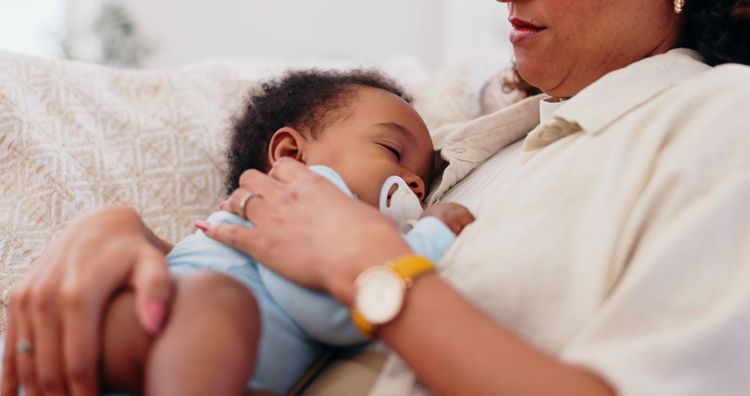 Eine Frau sitzt auf einem Sofa und hat ein Baby in einem Strampler auf der Brust, das schläft