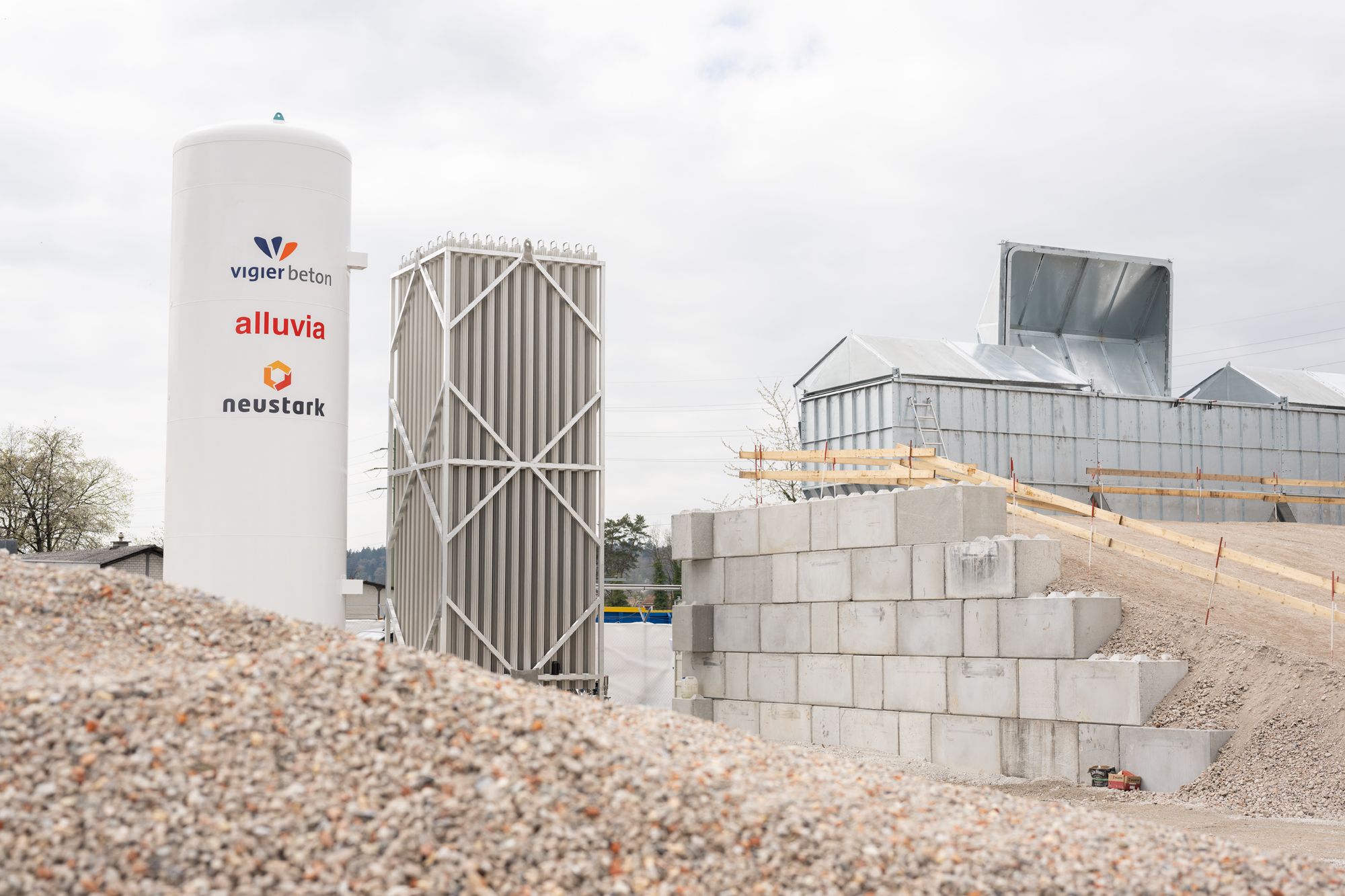 Anlage von neustark in Biberist, Solothurn, mit CO₂-Speichertank und Recyclingmaterial im Vordergrund; auf dem Tank sind die Logos von Vigier Beton, alluvia und neustark zu sehen.