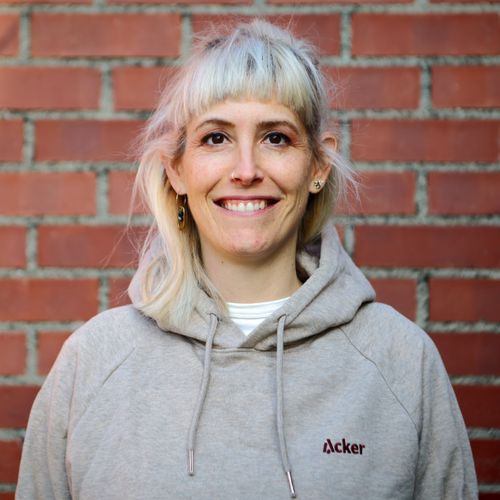Porträtaufnahme von Simone Nägeli. Sie lächelt in die Kamera; grauer Hoodie mit «Acker»-Logo, Backsteinwand im Hintergrund.